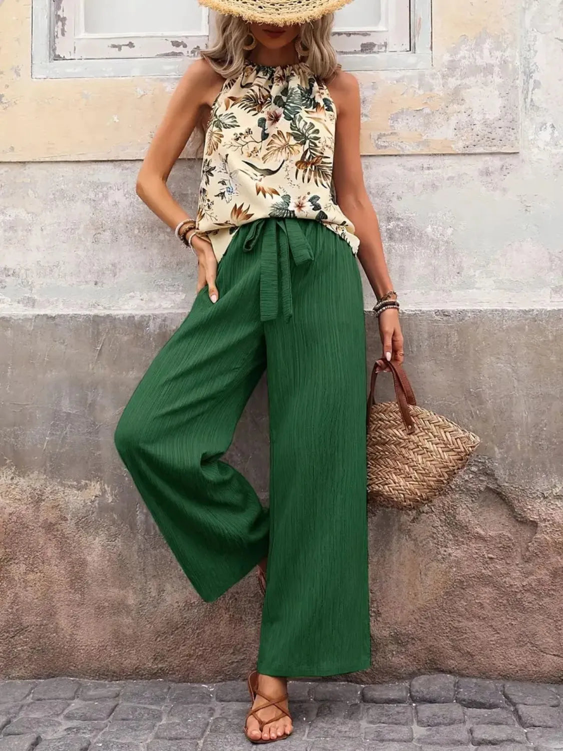 Woman wearing a printed tank top with tied neckline and wide leg green pants set, styled with a straw hat and wicker handbag.